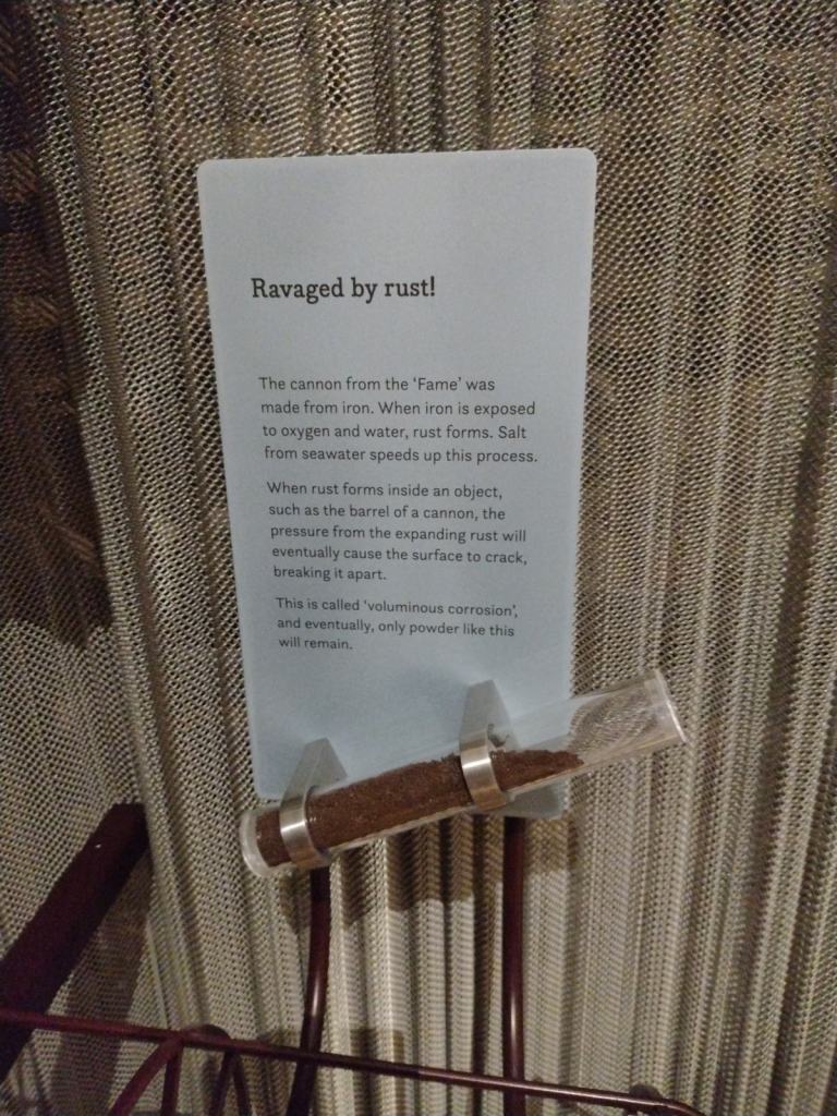 Ein Informationsschild über Rostschäden an einem Kanonenrohr, mit einem Probenglas, das rostiges Material enthält.